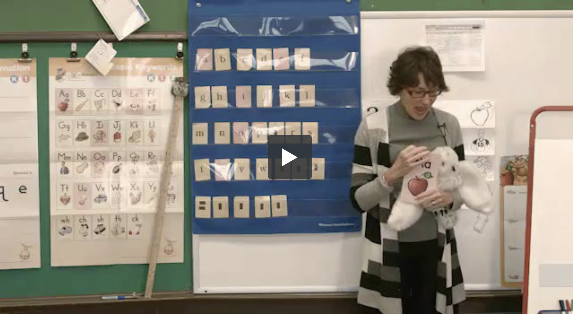 A teacher stands in a classroom holding a stuffed animal. Behind her, alphabet charts, a pocket chart with letter cards, and a green board are visible. The teacher is wearing a grey cardigan and glasses.